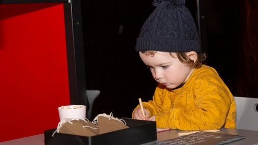 A young child with a yellow jumper and navy bobble hat sitting at a table writing a Christmas wish on a brown swing label